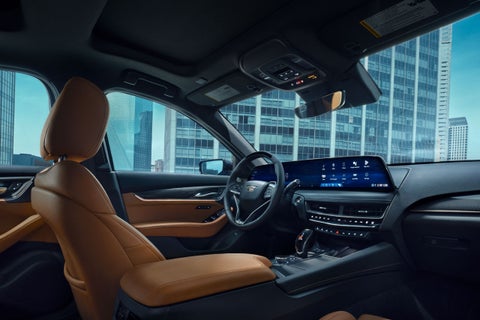 Interior view of a car with tan leather seats and a large digital dashboard, city buildings visible through windows.